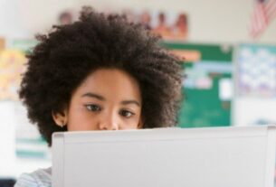 A imagem mostra uma criança com cabelos cacheados e volumosos, focada em um dispositivo eletrônico, possivelmente um laptop ou tablet branco, que está à sua frente. O fundo sugere um ambiente escolar: há um quadro verde e cartazes coloridos na parede.