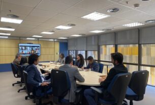 Reunião em uma sala de conferências, com algumas pessoas presentes na sala e outras participando por videochamada em uma tela grande.