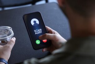 Um homem segura um smartphone que exibe um alerta de fraude ("SCAM ALERT") durante uma chamada recebida. Os botões para atender (verde) e recusar (vermelho) a chamada estão visíveis na parte inferior da tela. Na mão esquerda, ele segura um copo plástico transparente com gelo e um líquido claro, provavelmente água ou refrigerante. O fundo é desfocado, sugerindo um ambiente casual, como uma cafeteria, com uma mesa escura. A imagem ilustra o problema de chamadas fraudulentas e a importância de estar atento a alertas de segurança em dispositivos móveis.