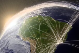 Uma imagem de satélite da Terra, focada na América do Sul, com linhas brancas brilhantes que representam conexões de rede ou rotas de voo irradiando-se sobre o continente e ao redor do globo. O sol está nascendo ou se pondo no horizonte, criando um brilho intenso no lado esquerdo da imagem.