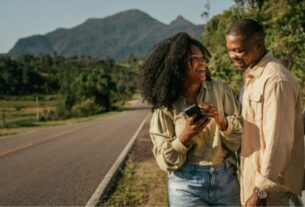 Na imagem, casal sorri ao lado do carro em uma estrada cercada por vegetação, e montanhas são visíveis no horizonte. Ambos sorriem enquanto a mulher segura um celular e o homem olha para o aparelho.
