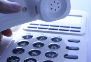 Uma mão segura o monofone de um telefone fixo branco, com o teclado numérico em foco.